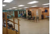 A spacious interior room with beige flooring and peach-colored walls, featuring a row of chairs along one wall, a staircase with wooden handrails on the left, and a small table with chairs near the back. The ceiling has multiple fluorescent lights, and there is a clock on the wall above a doorway.