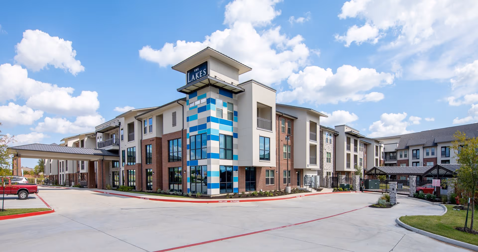 Exterior view of The Lakes at Grand Prairie senior living facility showing a modern three-story building with a mix of brick, stone, and blue accent panels under a partly cloudy sky. The driveway and parking area are visible in the foreground.