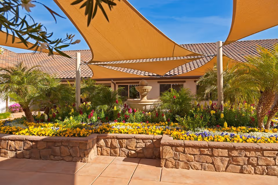Outdoor garden area with a stone raised flower bed filled with colorful flowers and palm plants, a central water fountain, and large beige shade sails overhead. The background shows a building with a tiled roof under a clear blue sky.