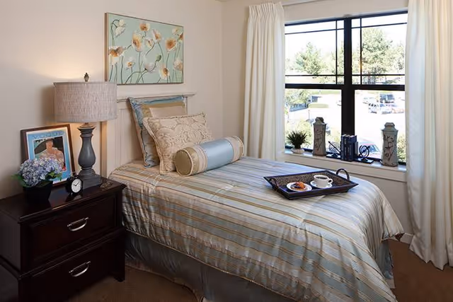 A neatly made single bed with striped bedding and decorative pillows in a bright bedroom. A dark wooden nightstand beside the bed holds a lamp, a small clock, a framed picture, and a potted plant. A large window with white curtains lets in natural light and shows a view of trees and parked cars outside. A tray with a cup of coffee and a pastry rests on the bed.