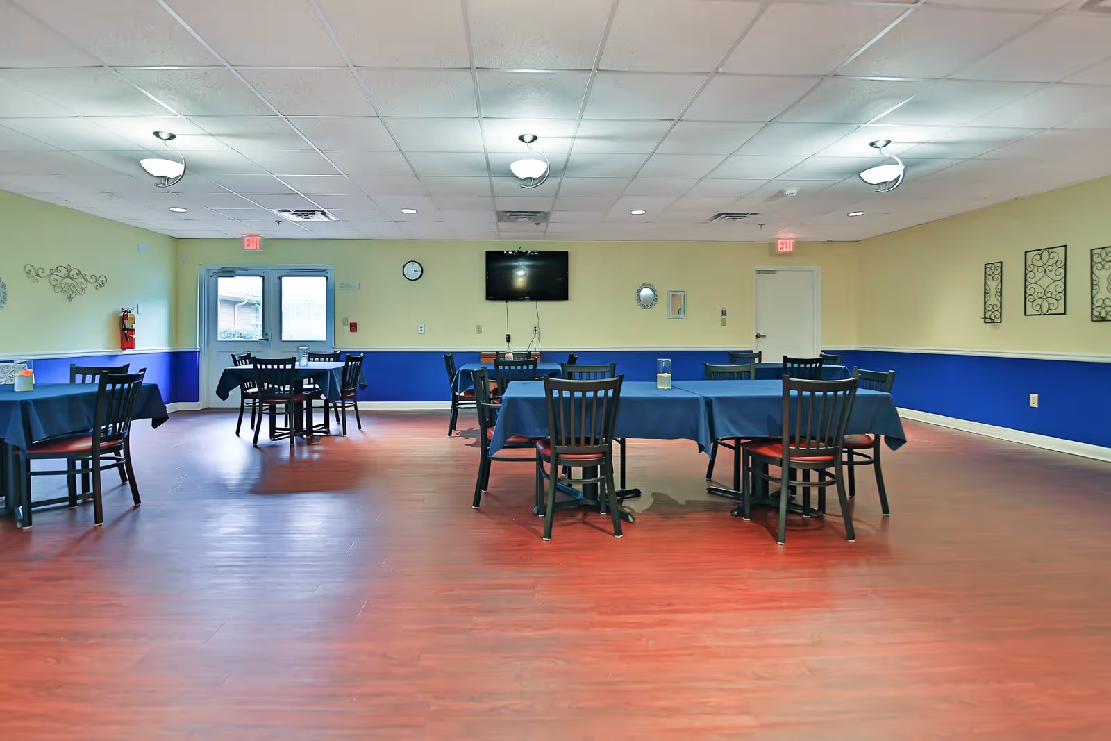 A spacious dining room with several tables covered in dark blue tablecloths and black chairs with red cushions. The room has light yellow walls with a blue lower half, wooden flooring, and ceiling lights. A wall-mounted TV, clock, and decorative mirrors are visible on the far wall, along with two exit doors.