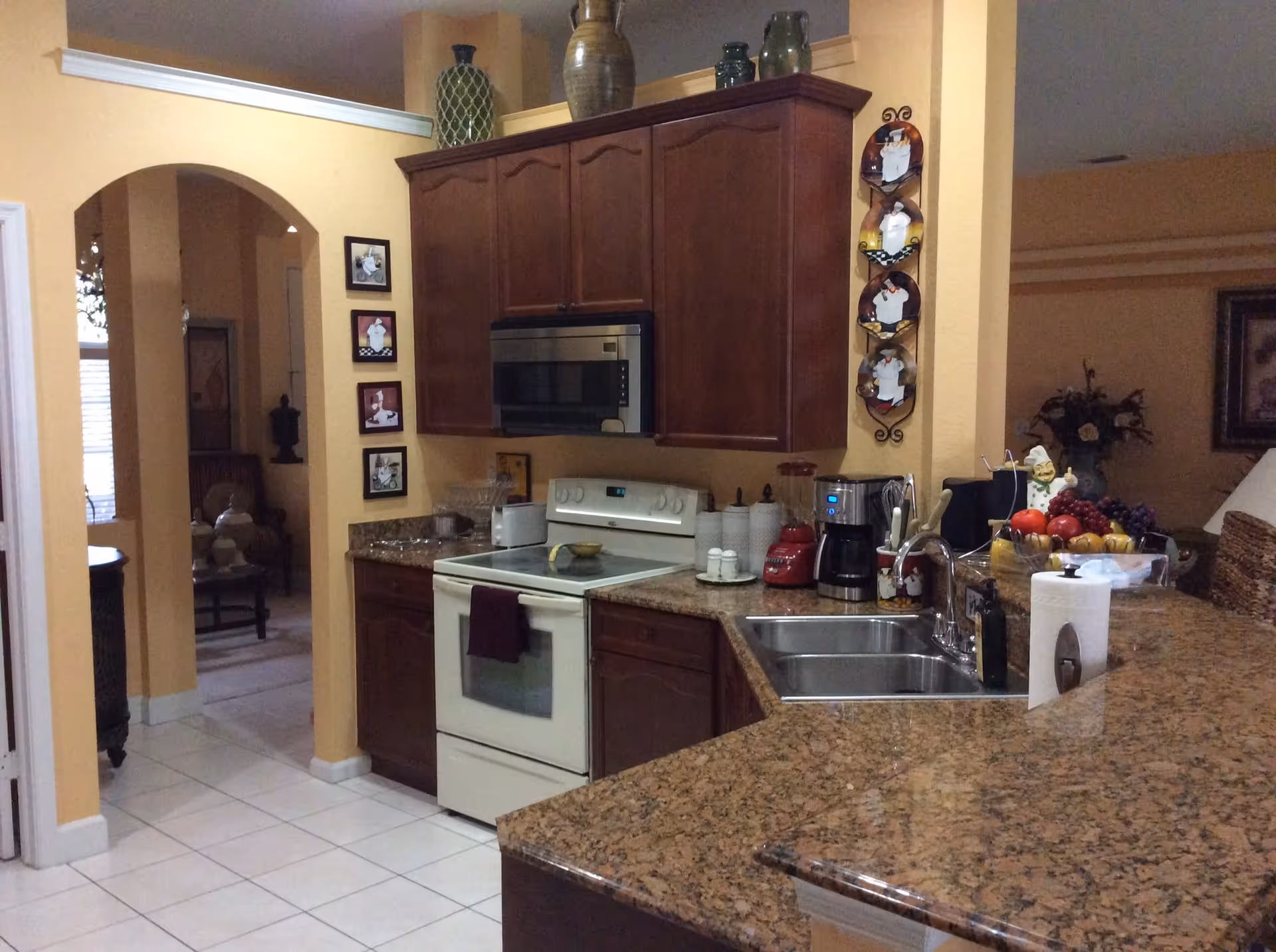 Open kitchen with granite countertops, wooden cabinets, a stove and microwave, double sink, and small appliances on the counter.