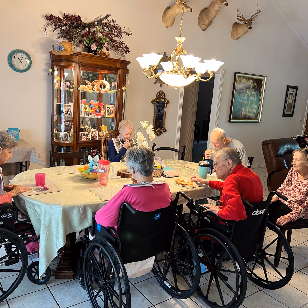 A group of elderly individuals, some in wheelchairs, seated around a dining table in a well-lit room. The table is covered with a light-colored tablecloth and has various items on it including cups, a basket, and a small stuffed bunny. Behind them is a wooden display cabinet with decorative items and photos, a wall clock, and mounted deer heads on the wall. A chandelier hangs above the table providing warm light.