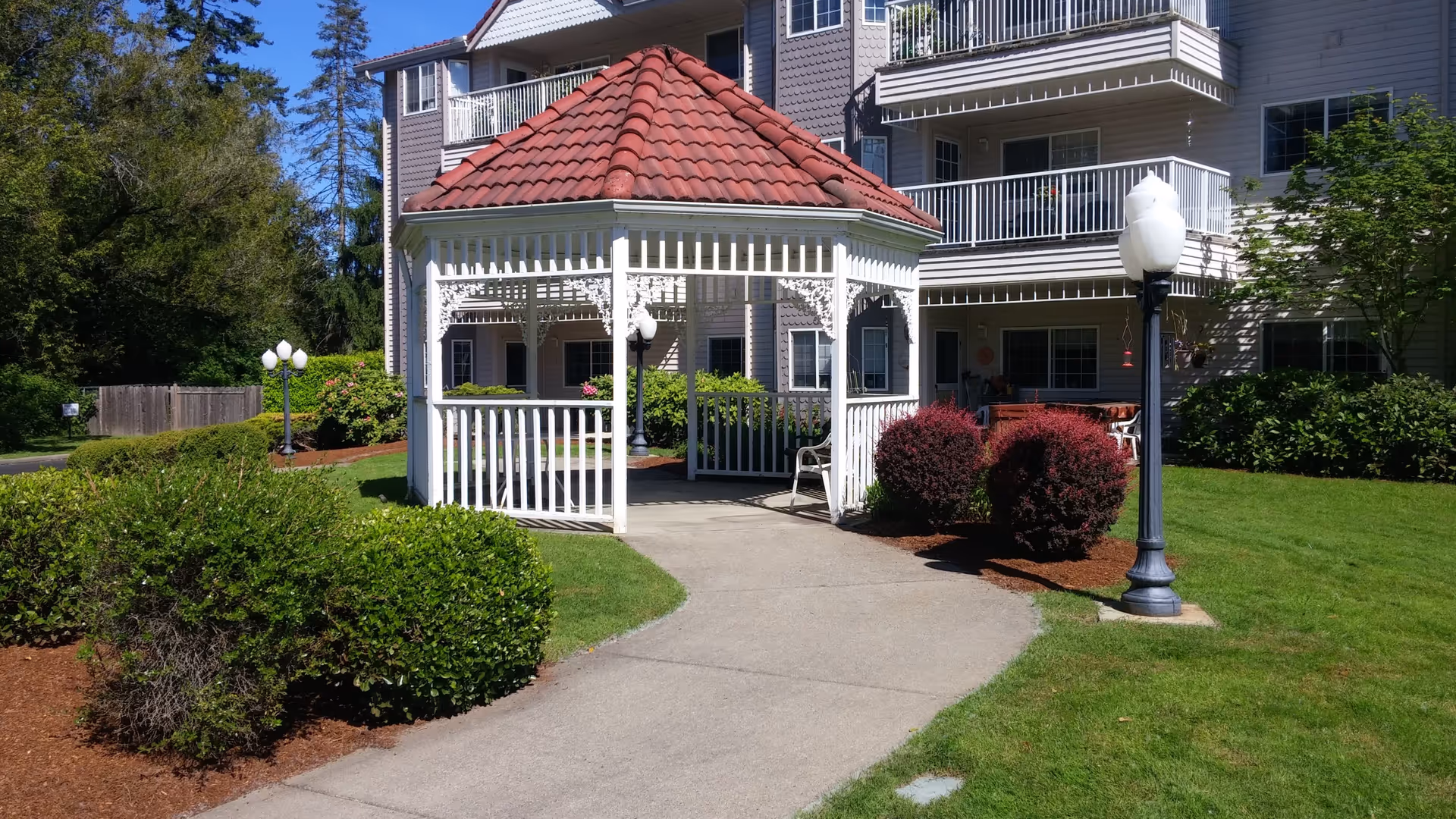 Outdoor view of a senior living facility showing a white gazebo with a red tiled roof on a paved pathway surrounded by green grass, bushes, and trees. The building in the background has balconies and windows, with lamp posts near the gazebo.