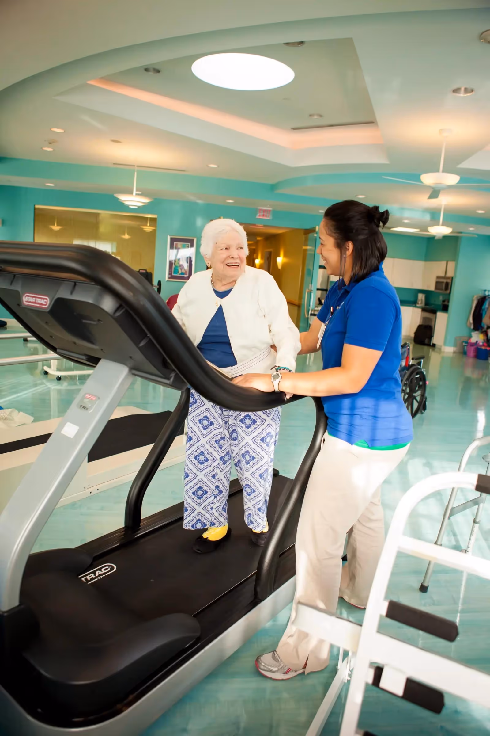 An elderly woman standing on a treadmill smiling and holding the handrails while a caregiver in a blue shirt supports her and smiles back in a bright, spacious rehabilitation or exercise room.