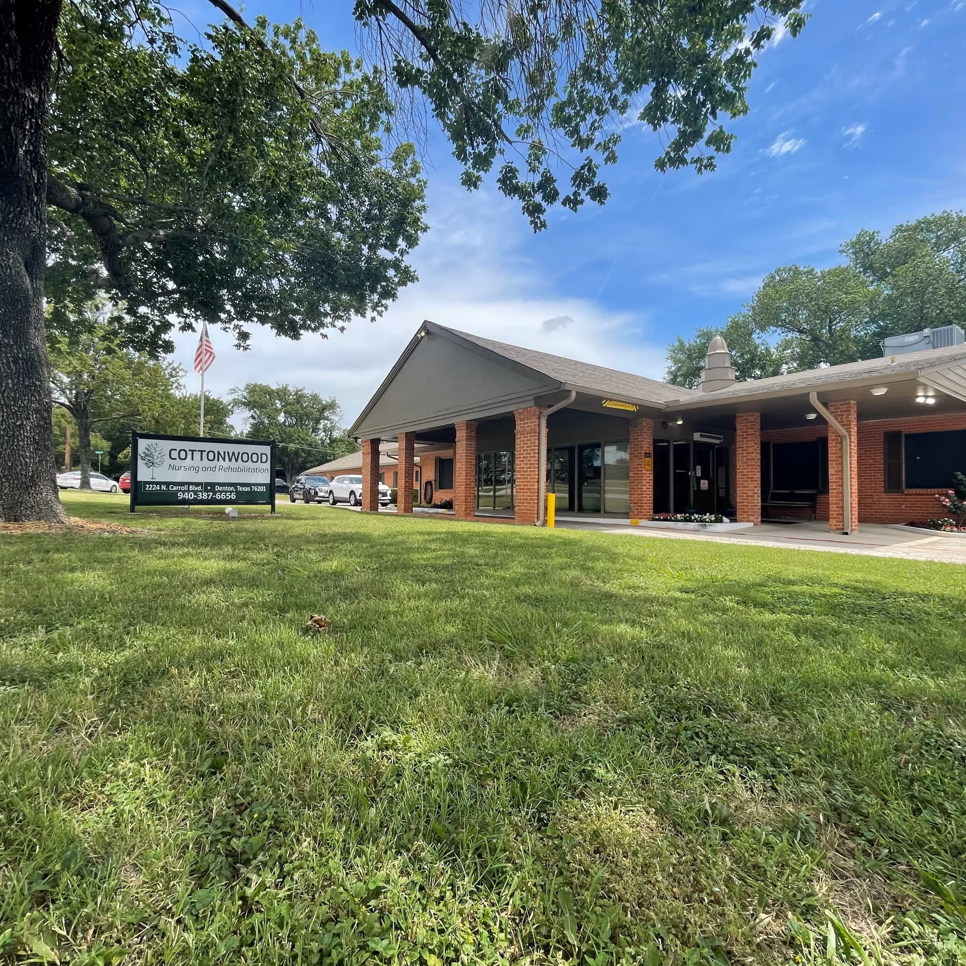 Exterior view of Cottonwood Nursing and Rehabilitation facility with a brick building, a covered entrance, green lawn, large tree, and a sign displaying the facility name, address, and phone number under a partly cloudy sky.