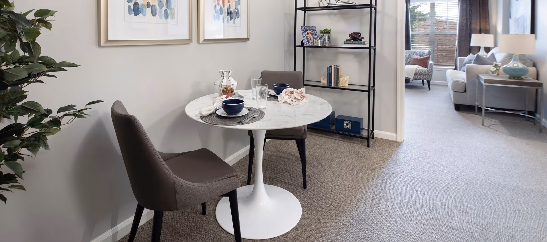 Small dining nook with a round white table and two gray chairs beside shelving and a view into a living room with a sofa.