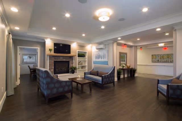 A spacious and well-lit senior living common area with wooden flooring, two blue patterned sofas, a matching armchair, a wooden coffee table, a stone fireplace with a TV mounted above it, built-in shelves with decorative items, and framed artwork on the walls.