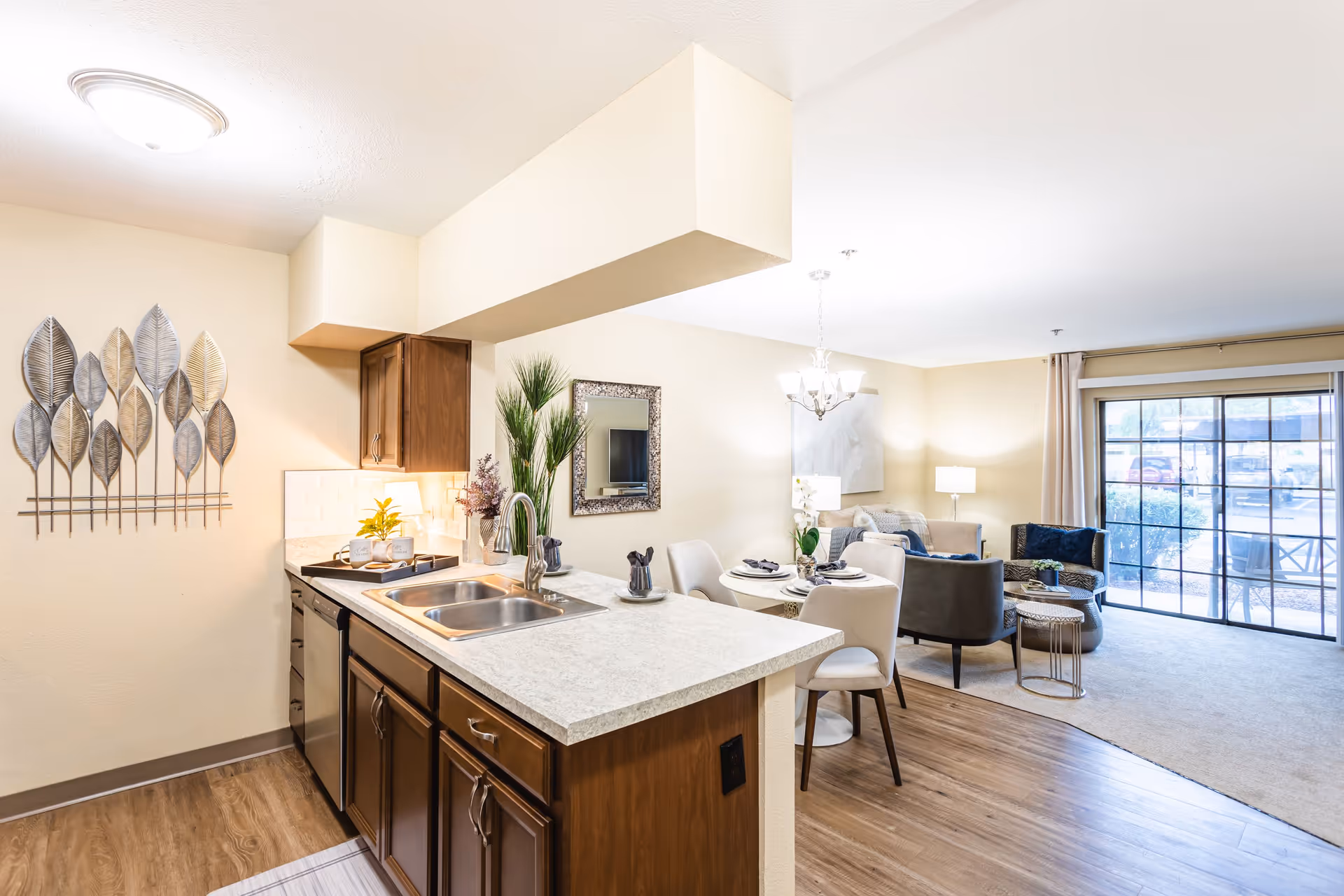 Bright and modern open-concept living space featuring a kitchen with a double sink and wooden cabinets, a small dining area with a round table set for four, and a cozy living room with armchairs and a sofa near large sliding glass doors leading to an outdoor patio.