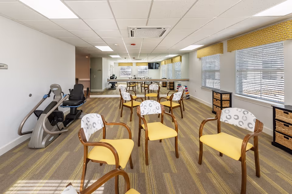 A bright room with several wooden chairs featuring yellow seats and patterned backs arranged in a circle on a carpeted floor. The room has large windows with blinds and yellow valances, a mirrored wall, and exercise equipment including a recumbent bike. There are also small storage units with wicker drawers along the walls.