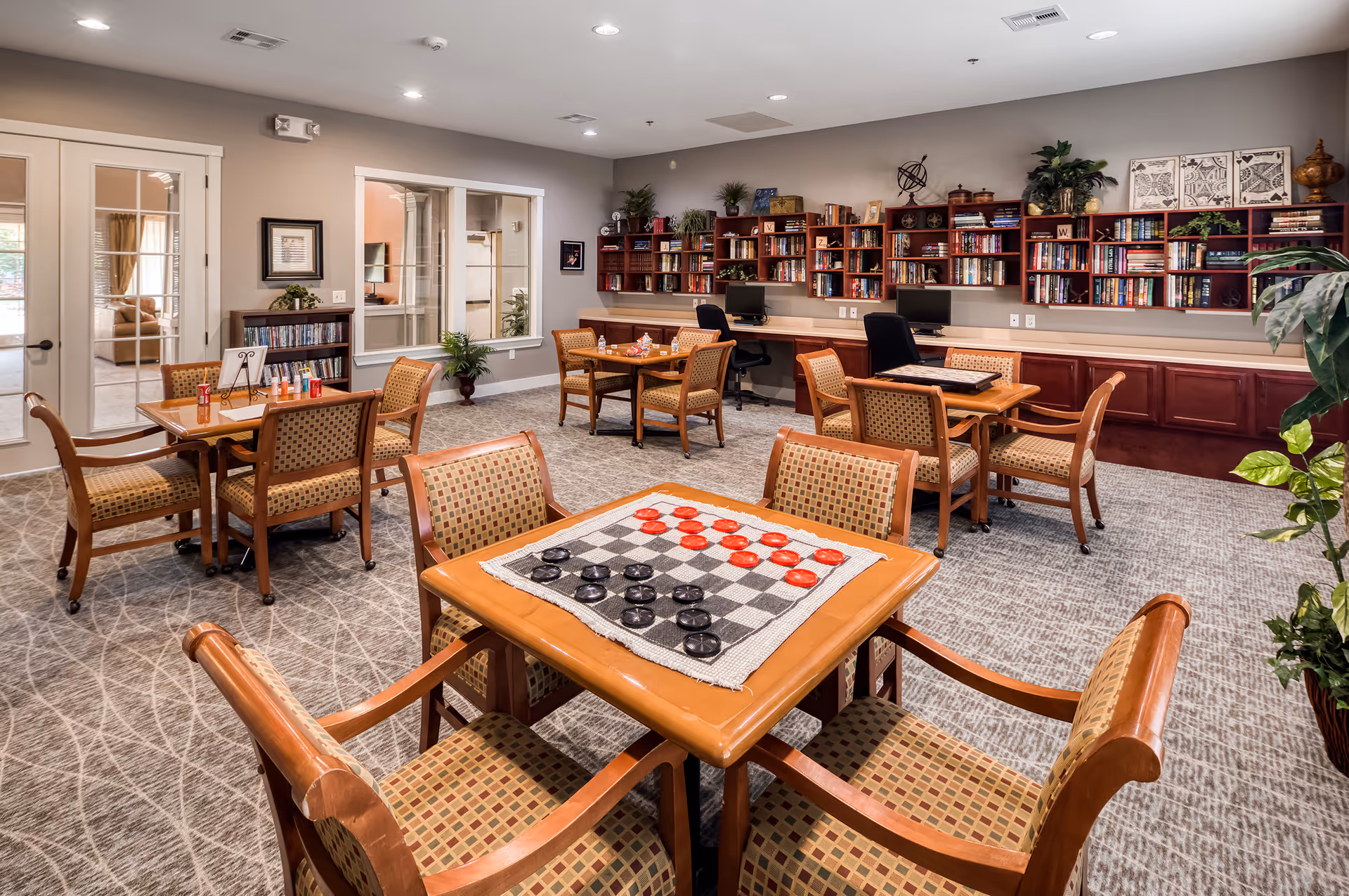 Bright assisted-living common room with tables and chairs, a checkers board on the foreground table, and bookshelves and computers along the back wall.