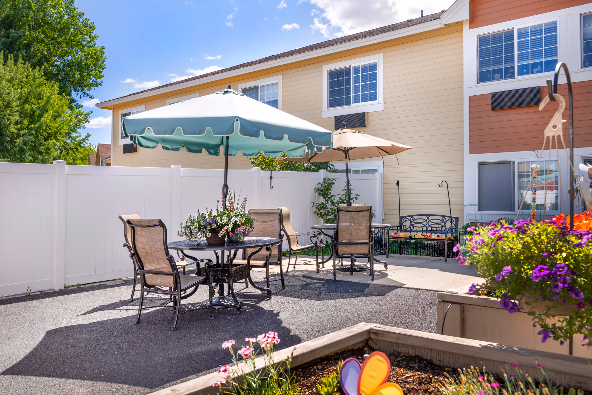 Sunlit outdoor patio with umbrella-shaded tables and chairs, flower planters, and a two-story building facade in the background.