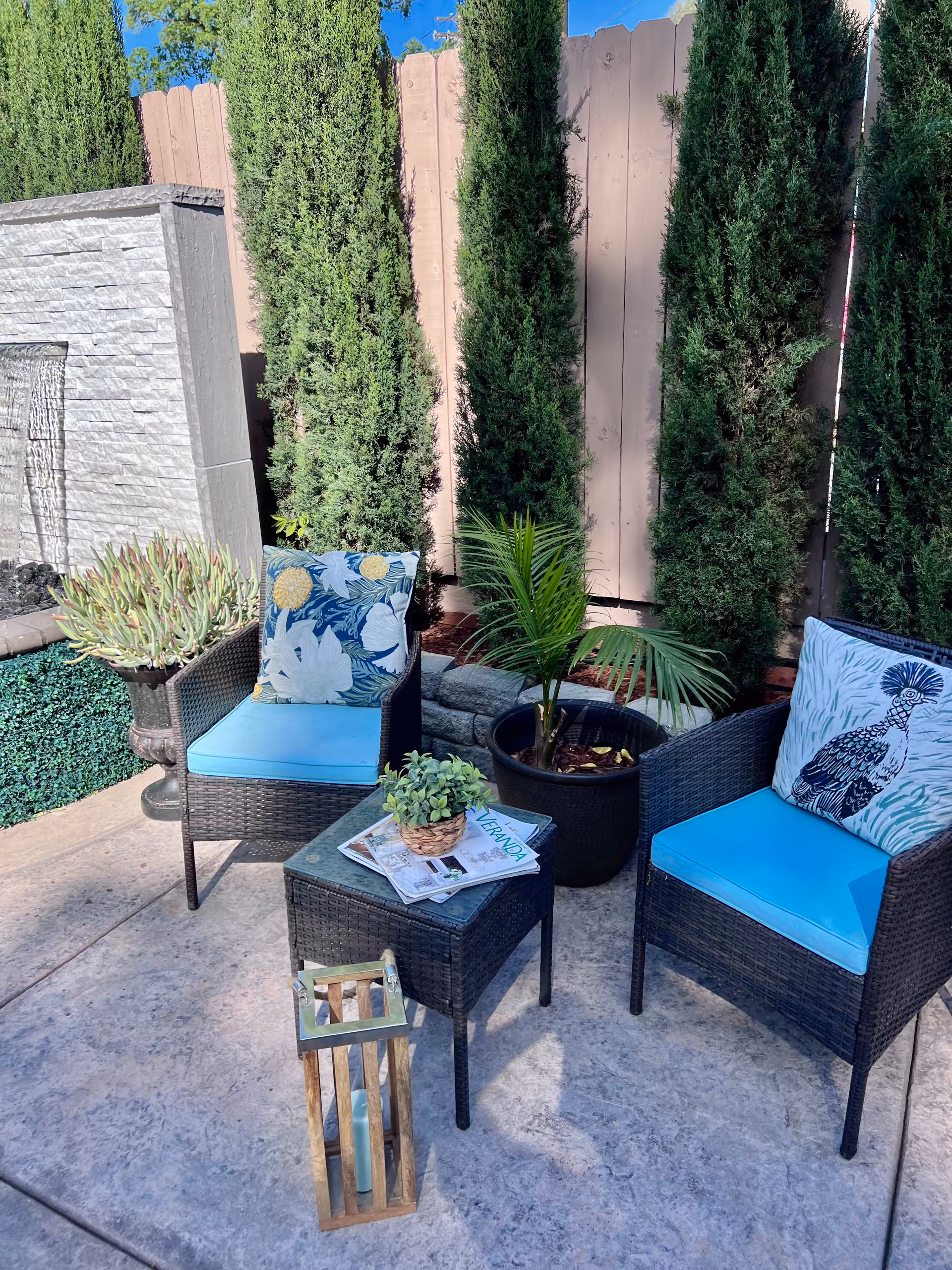 Outdoor patio area with two wicker chairs featuring blue cushions and decorative pillows, a small wicker table with magazines and a small potted plant, a wooden lantern on the ground, tall green trees, and a stone water feature against a wooden fence.
