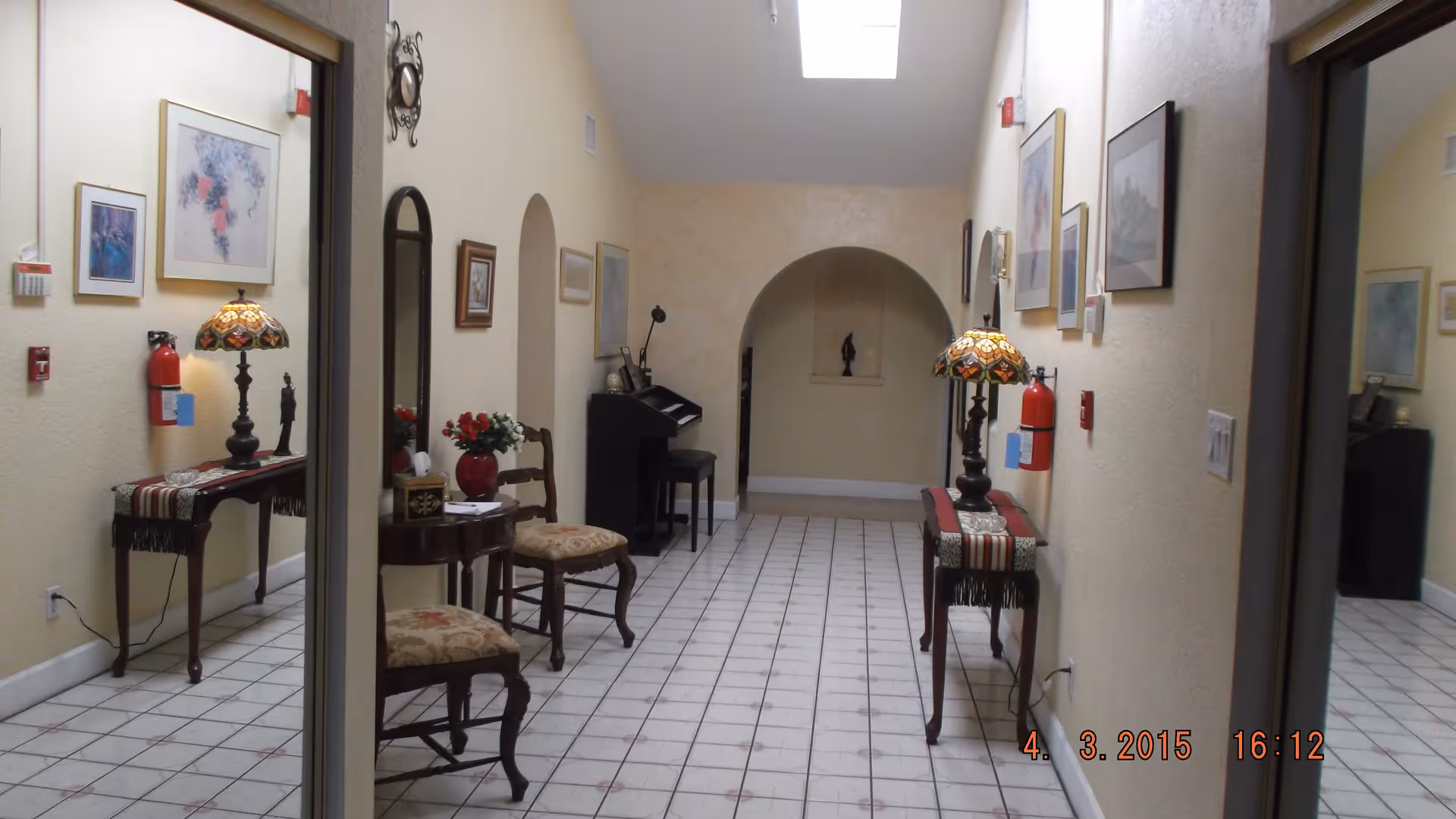 Tiled hallway interior with chairs, small tables and lamps, framed artwork, and a piano in an arched alcove.