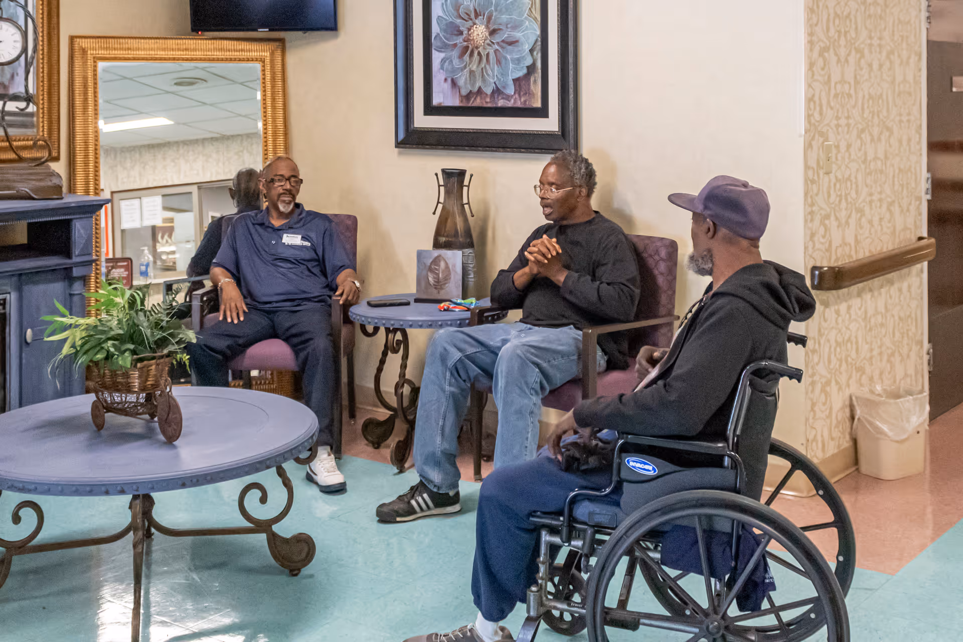 Three elderly men sitting and conversing in a common area of a senior living facility. One man is in a wheelchair, while the other two are seated in chairs around a small table. The room has light-colored walls, a decorative vase, a framed flower picture, and a round coffee table with a plant on it.