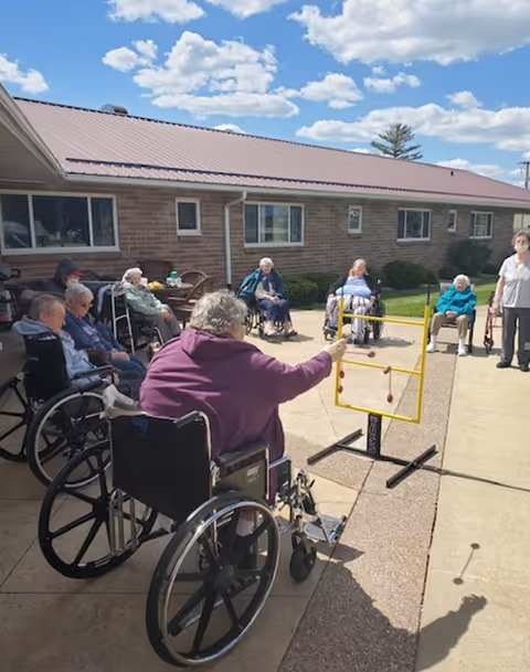 A group of elderly people, most in wheelchairs, are gathered outside on a sunny day at a convalescent home. They are playing a ring toss game on a paved patio area in front of a single-story brick building with a metal roof. The sky is blue with scattered clouds.