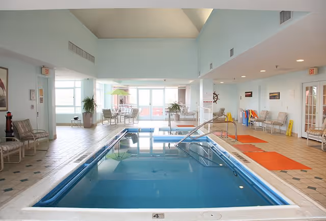 Indoor swimming pool area with clear blue water, surrounded by tiled flooring and several chairs along the walls. There are tables with umbrellas and plants near large windows letting in natural light. The ceiling is high with recessed lighting.