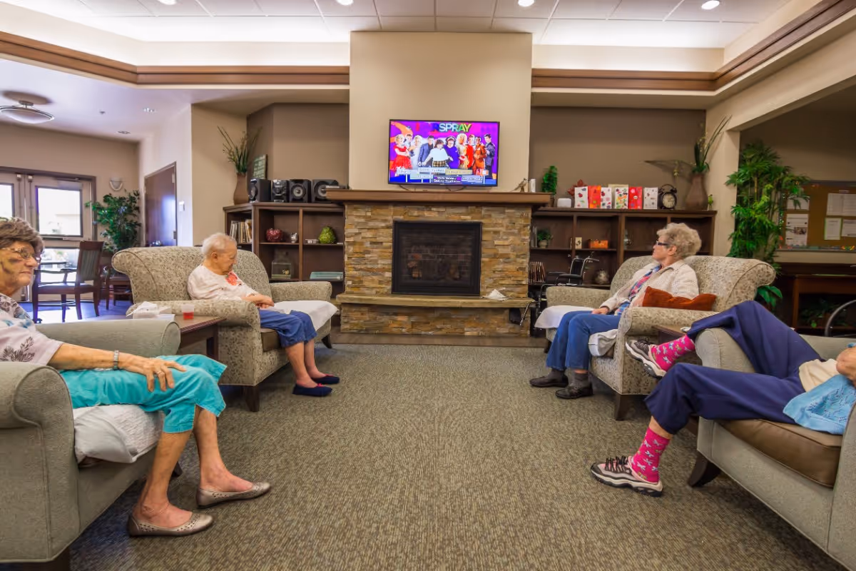 A cozy living room in a senior living facility with four elderly women seated on armchairs and sofas arranged around a stone fireplace. A flat-screen TV is mounted above the fireplace, and shelves with decorative items and plants flank the fireplace. The room has warm lighting and a carpeted floor.