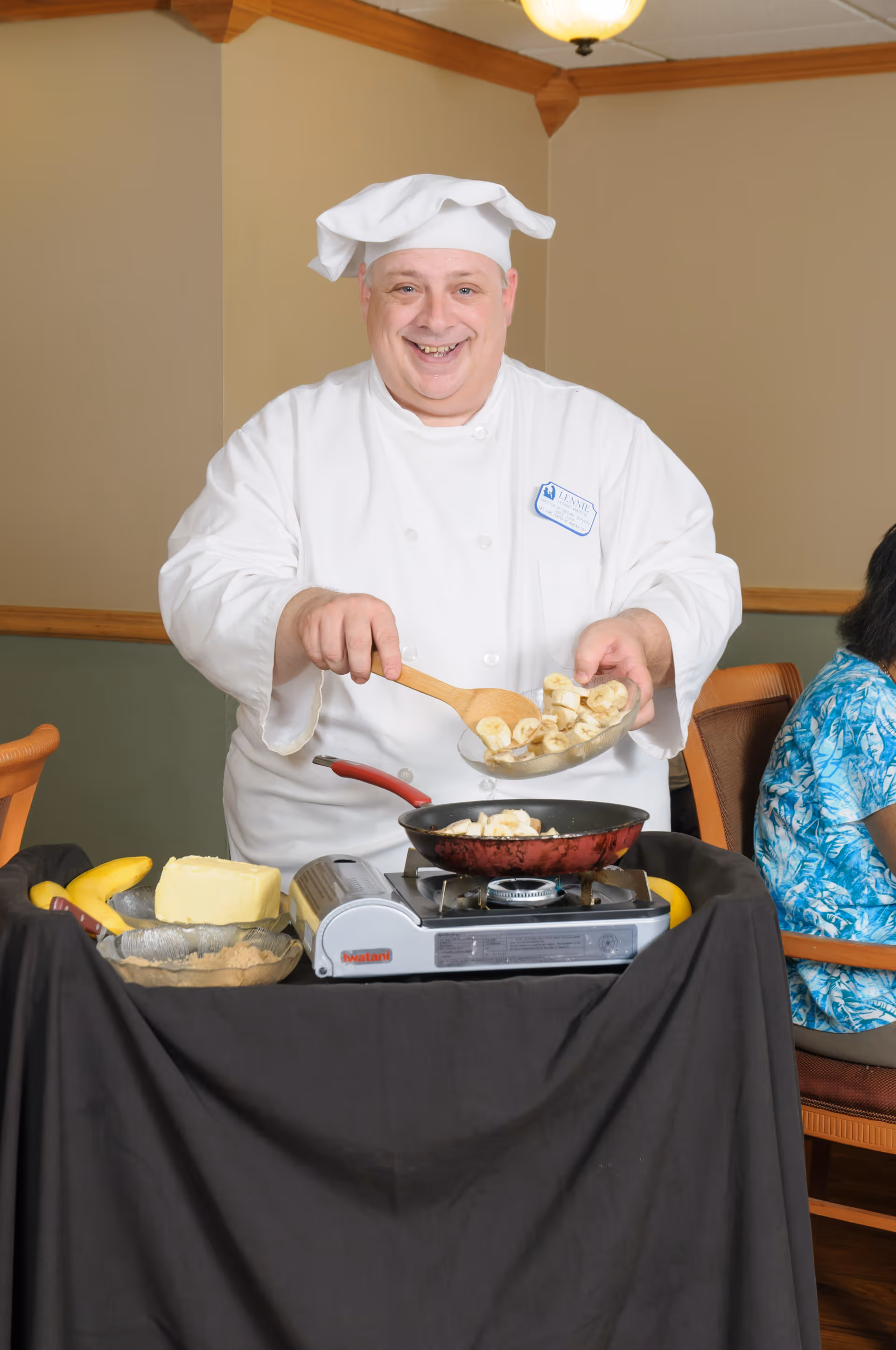 A smiling chef wearing a white chef's hat and coat is cooking sliced bananas in a red frying pan on a portable stove. There are bananas and a block of butter on the table covered with a black cloth. A person in a blue patterned shirt is seated in the background.