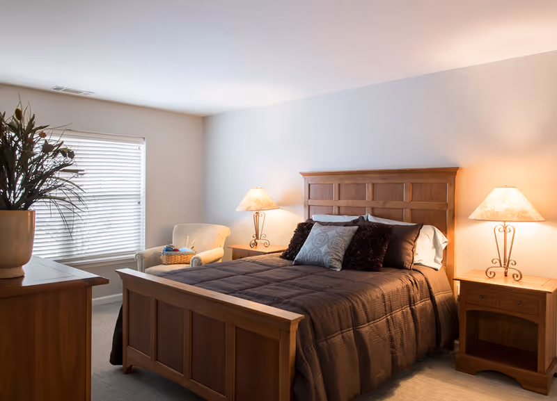A cozy bedroom with a wooden bed frame and headboard, a dark brown comforter, and several decorative pillows. There are two matching wooden nightstands on either side of the bed, each with a lamp emitting warm light. A comfortable armchair with a basket on it is placed near a window with closed blinds. A wooden dresser with a large potted plant is visible in the foreground.