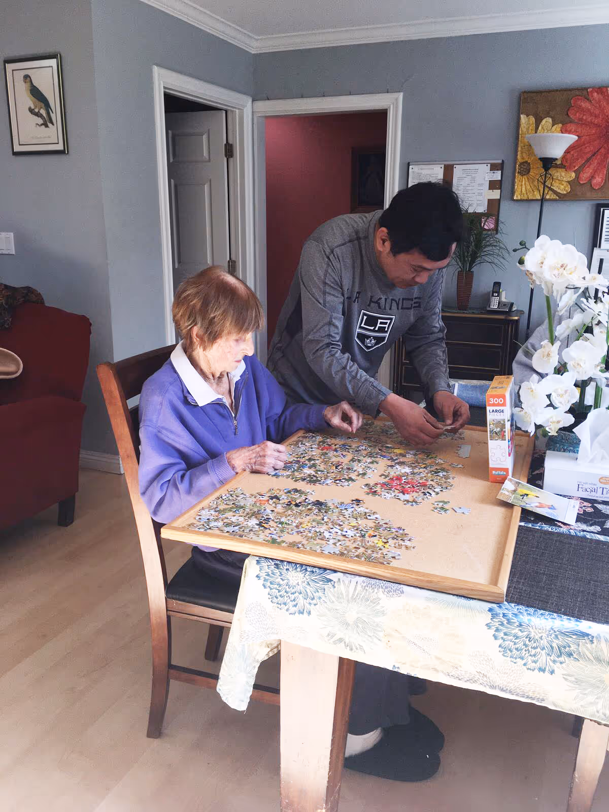 An elderly woman and a man are sitting at a table working on a jigsaw puzzle together in a cozy room. The table is covered with a floral tablecloth and puzzle pieces are spread out on a board. The room has light-colored walls, a red armchair, a framed bird picture, a floor lamp, and a vase with white flowers on the table.