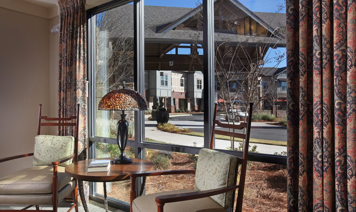 Two cushioned wooden chairs with a small round wooden table between them, holding a decorative lamp and a book, positioned next to large windows with patterned curtains, overlooking the outdoor area of a senior living facility.