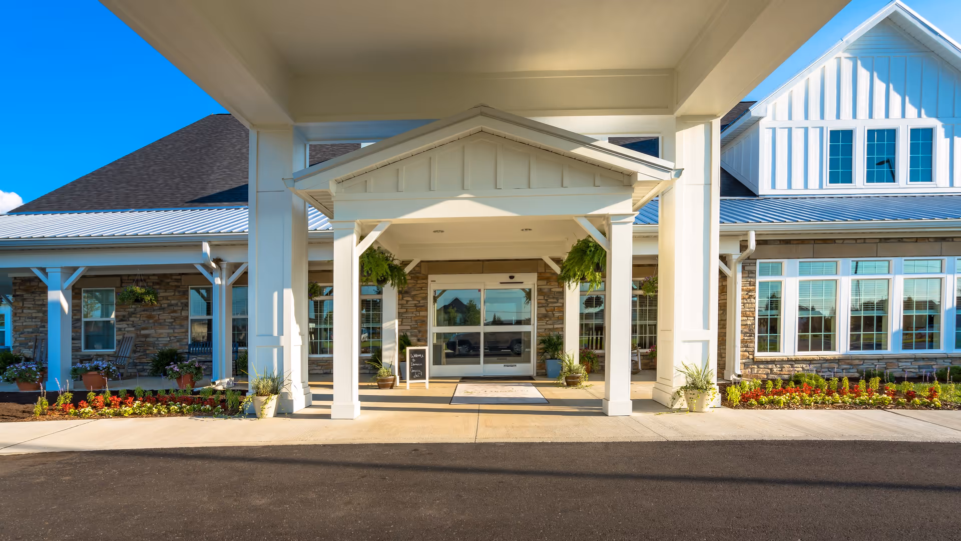 Front entrance of Demaree Crossing facility with a covered driveway, white pillars, hanging plants, and a welcoming mat. The building features large windows and stone accents under a clear blue sky.