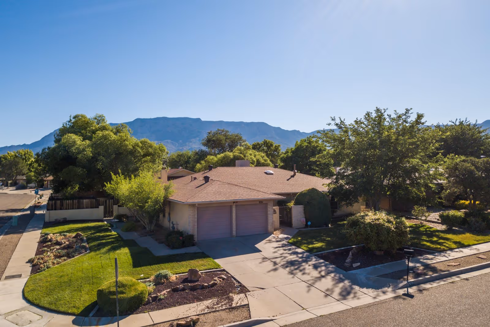 A single-story residential building with a two-car garage surrounded by green lawns, trees, and shrubs. The building is situated on a corner lot with a paved driveway and sidewalks. In the background, there are mountains under a clear blue sky.