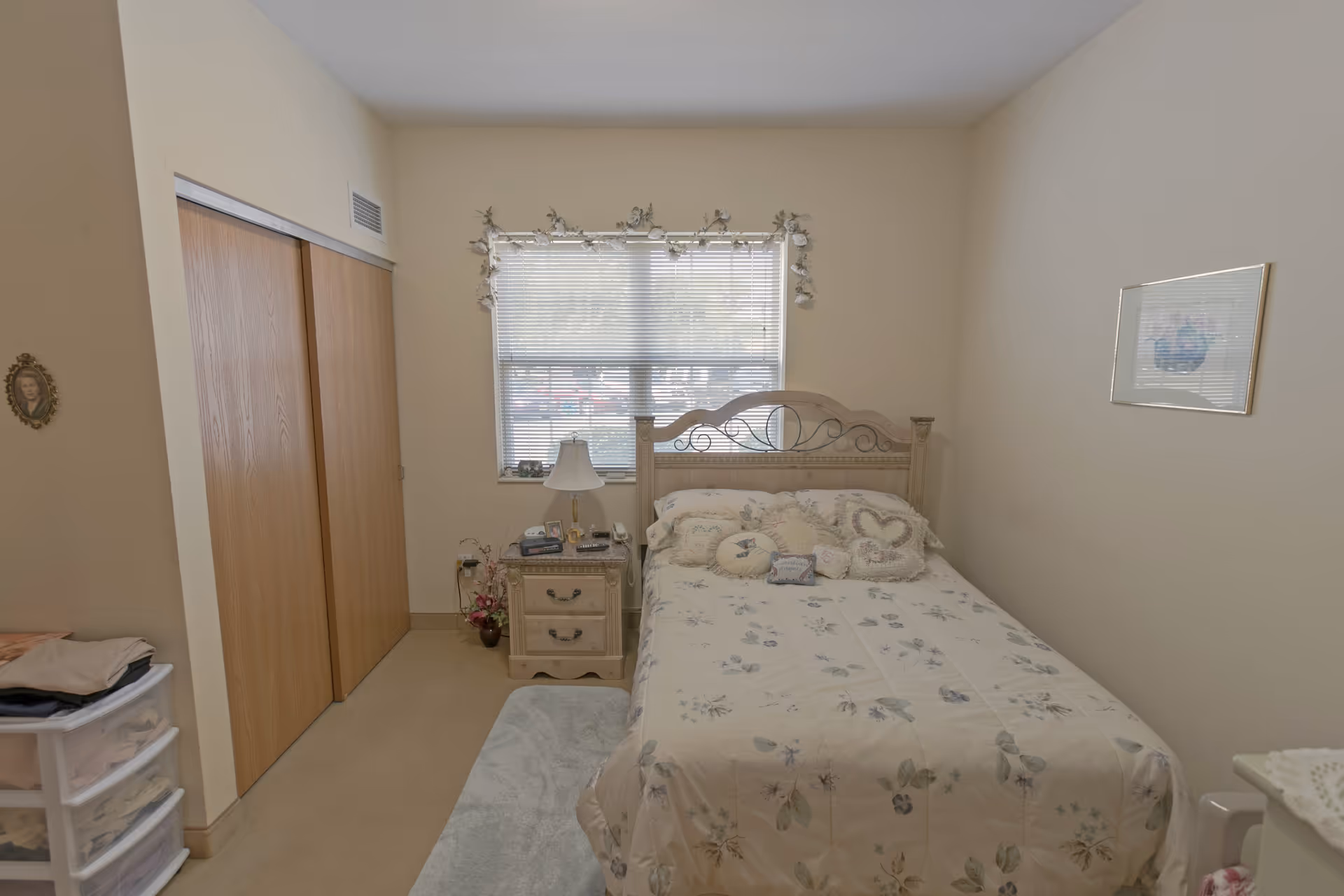 A small bedroom with a bed covered in floral bedding and multiple decorative pillows. There is a wooden nightstand with a lamp and various items next to the bed. A window with blinds and floral garland is centered on the wall behind the bed. To the left, there is a closet with wooden sliding doors. The walls are painted a light beige color, and a framed picture hangs on the right wall.