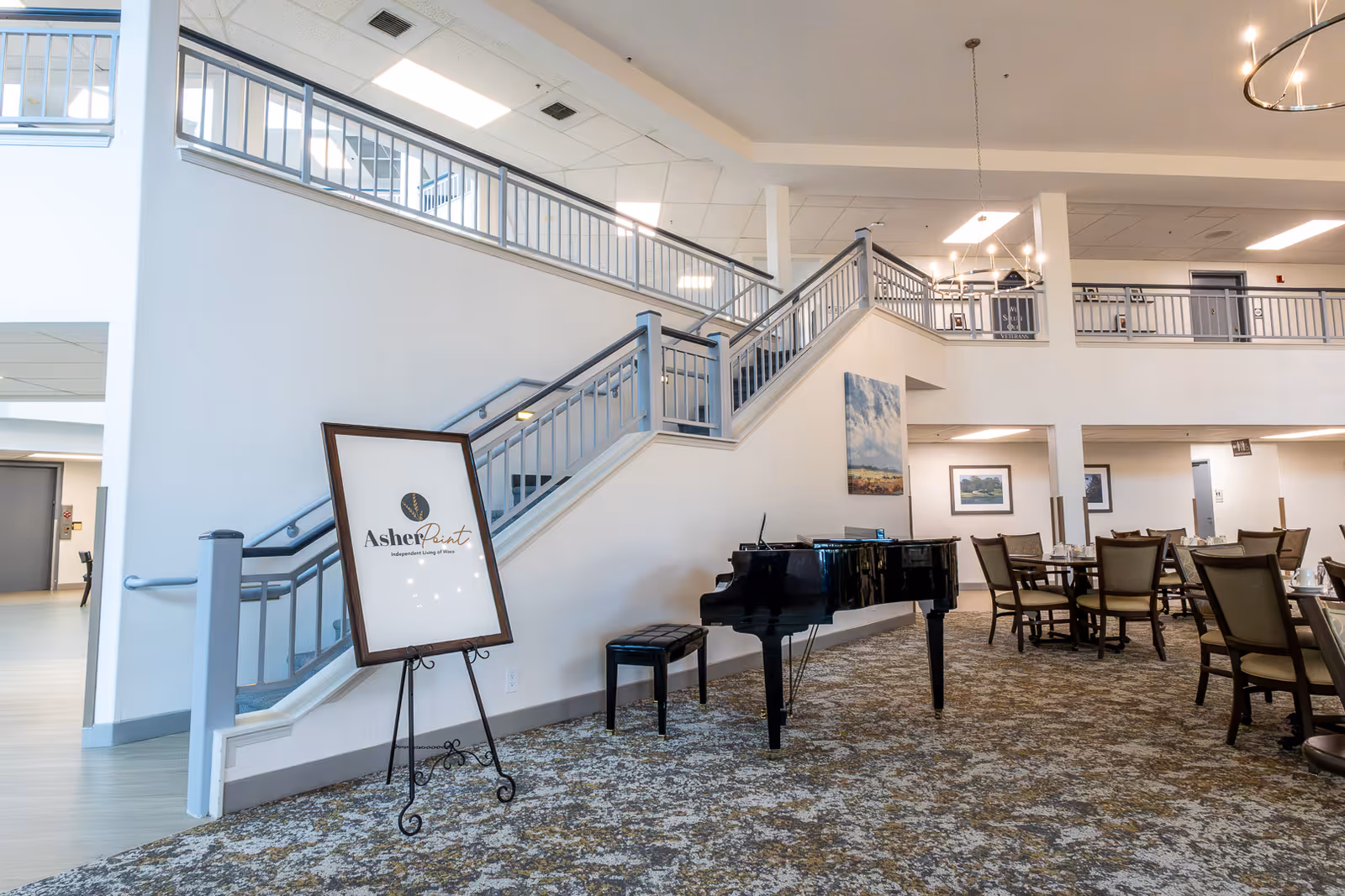 Spacious two-story common area in a senior living facility featuring a grand piano, dining tables, and a central staircase.