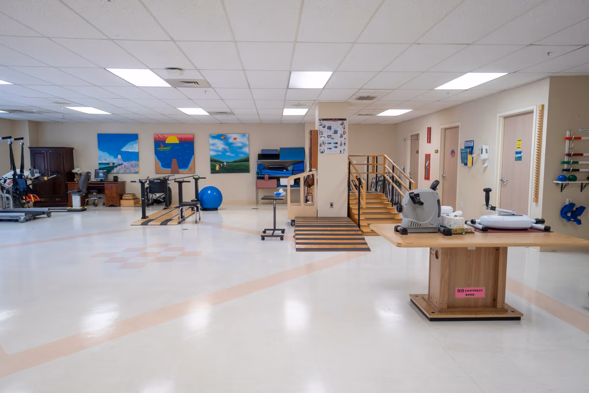 A spacious physical therapy or rehabilitation room with exercise equipment including parallel bars, a stationary bike, a treadmill, and various therapy tools. There are colorful paintings on the far wall and a wooden ramp and stairs in the center. The room has a clean, bright appearance with a tiled floor and fluorescent ceiling lights.