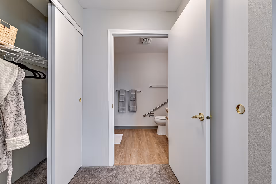 View through an open door into a bathroom with a toilet, grab bar, and two gray towels hanging on a towel rack. In the foreground, there is a closet with sliding doors, one of which is open showing a hanging robe and some hangers.
