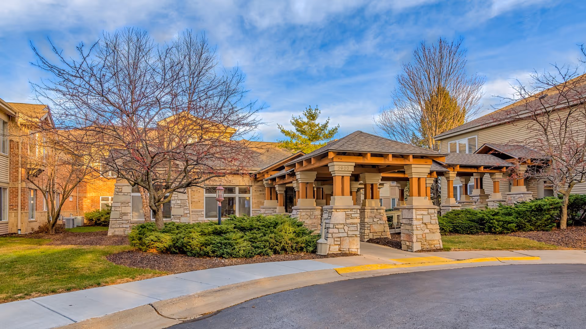 Exterior view of Harbour Health Assisted Living facility showing a covered entrance with stone pillars and wooden beams, surrounded by landscaped bushes and leafless trees under a partly cloudy blue sky.