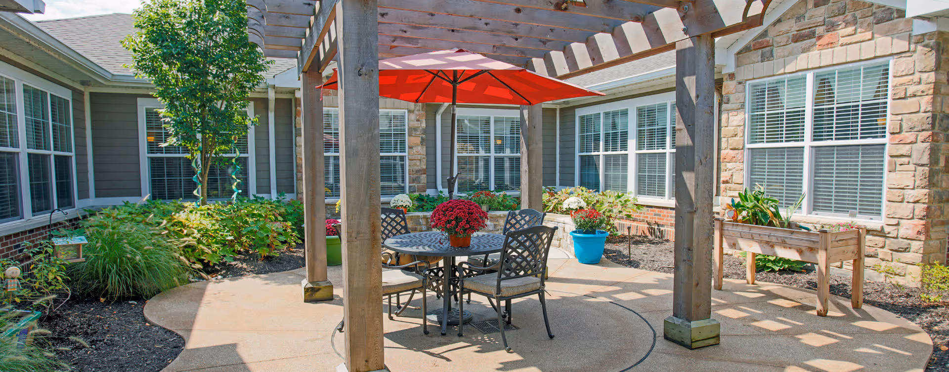 Outdoor patio area with a wooden pergola, a round metal table with four chairs, and a red umbrella providing shade. The patio is surrounded by garden beds with various plants and flowers, and the building's windows and stone exterior walls are visible in the background.