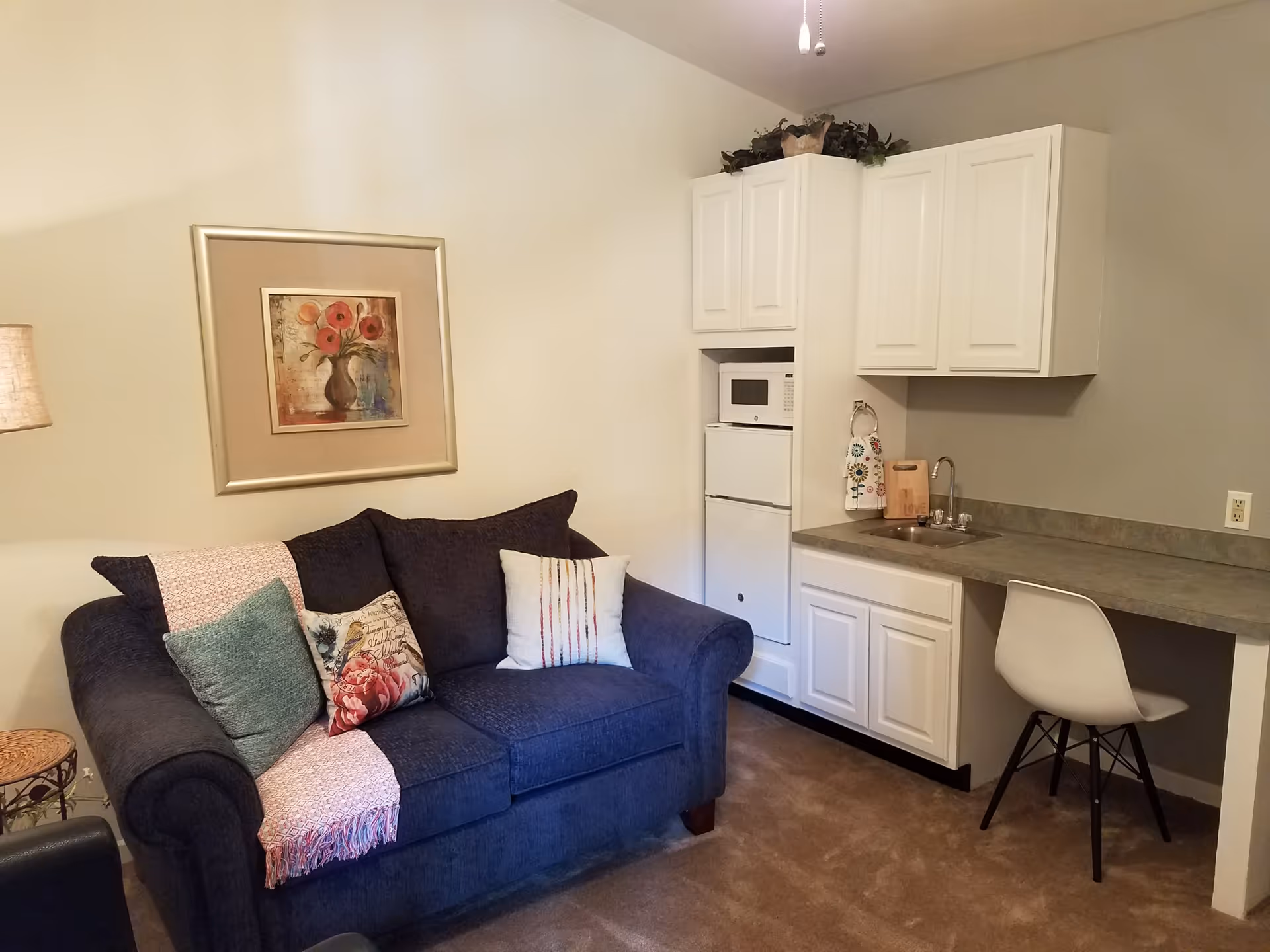 A cozy living area with a dark blue loveseat adorned with three decorative pillows and a pink throw blanket. On the wall above the loveseat is a framed painting of red flowers in a vase. To the right is a small kitchenette with white cabinets, a microwave, a mini refrigerator, a sink, and a white chair tucked under the counter.