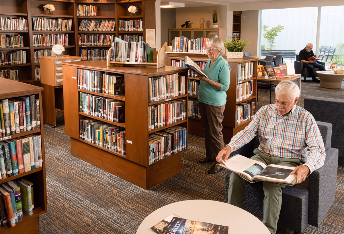 A cozy library area in a senior living facility with wooden bookshelves filled with books. An elderly woman stands reading a book while an elderly man sits in a gray armchair looking at a large book. In the background, another elderly man is seated near a window reading. The room is well-lit with natural light and has a carpeted floor.