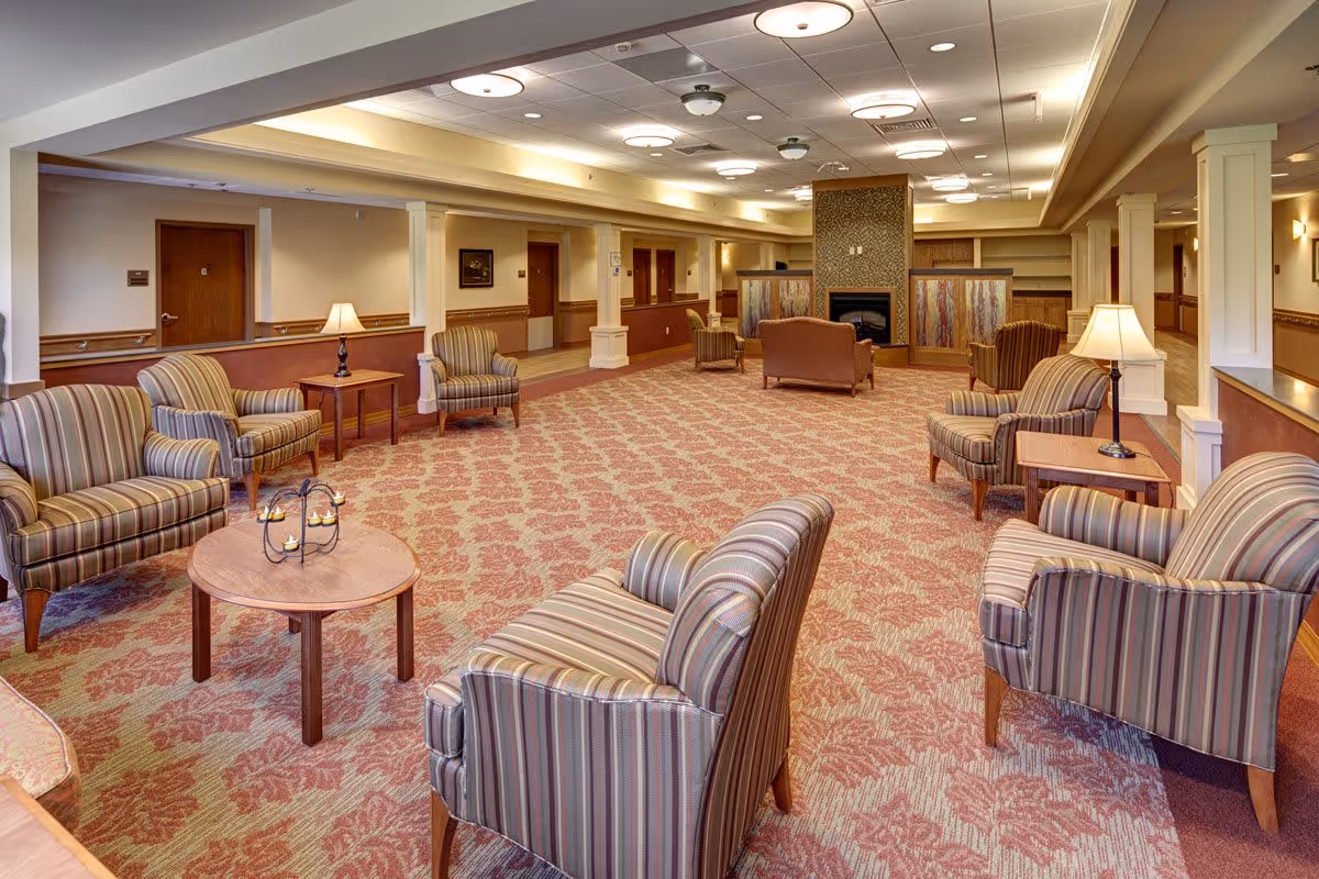 Spacious common lounge in an assisted living facility with multiple striped armchairs, side tables, lamps, and a fireplace at the far end.