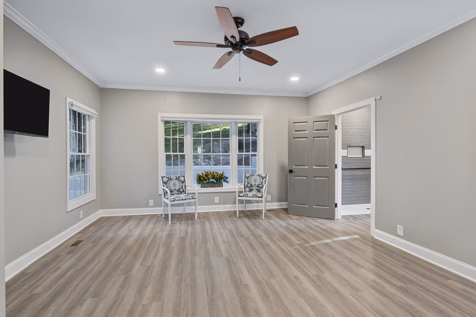 A spacious, empty room with light wood flooring, gray walls, and white trim. Two patterned chairs with a small table holding a flower arrangement are placed in front of a large window. A ceiling fan with wooden blades is mounted on the white ceiling, and a flat-screen TV is mounted on the left wall. An open gray door leads to another room with gray tiled walls.