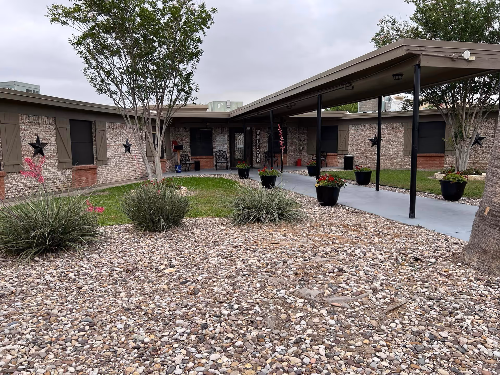 Front exterior of a one-story nursing facility with a covered entry walkway, potted plants, trees, and rock landscaping.