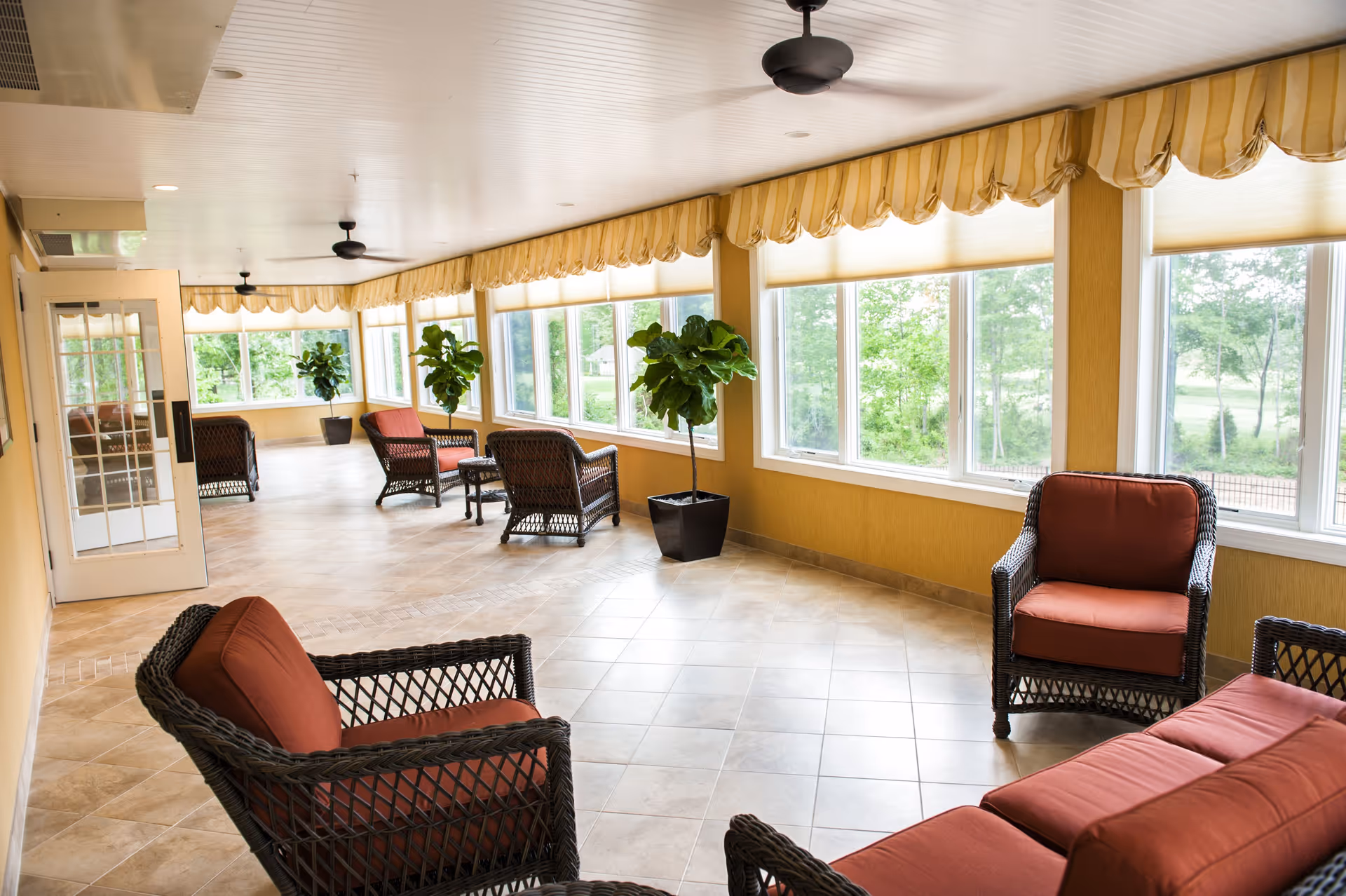 A bright sunroom with large windows along one wall, letting in natural light and offering a view of green trees outside. The room features several wicker chairs and sofas with rust-colored cushions arranged in small groupings. There are potted plants placed near the windows and ceiling fans overhead. The floor is tiled with a light beige color, and the walls are painted a warm yellow. The windows have yellow and white striped valances.