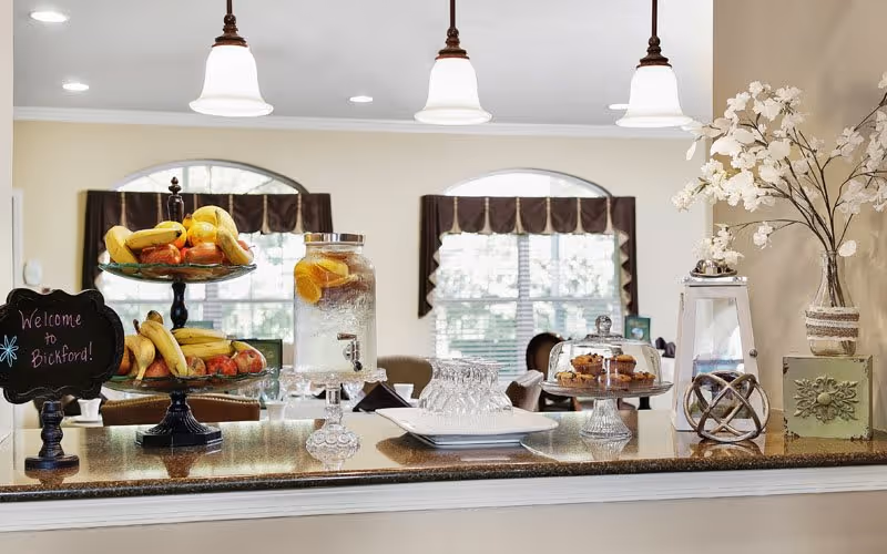 A countertop in a dining or common area with a tiered fruit stand holding bananas and apples, a glass dispenser filled with water and lemon slices, a plate with glasses, a covered plate with muffins, and decorative items including a vase with white flowers and a lantern. In the background, there are windows with dark valances and chairs around tables.