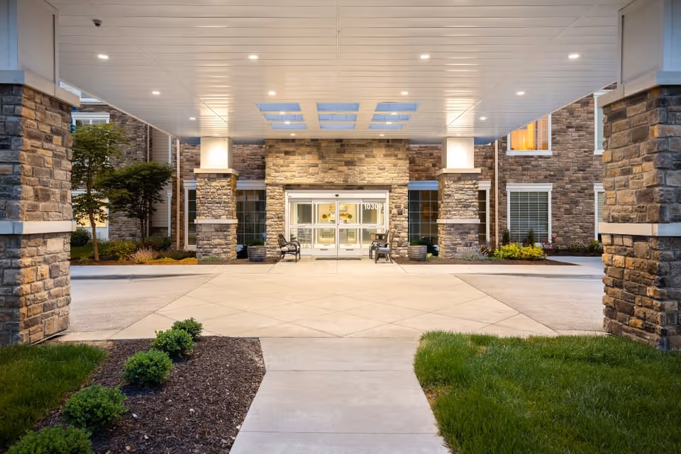 Entrance of a senior living facility with a covered drop-off area supported by stone pillars, glass double doors, benches on either side, and landscaped greenery around the walkway.