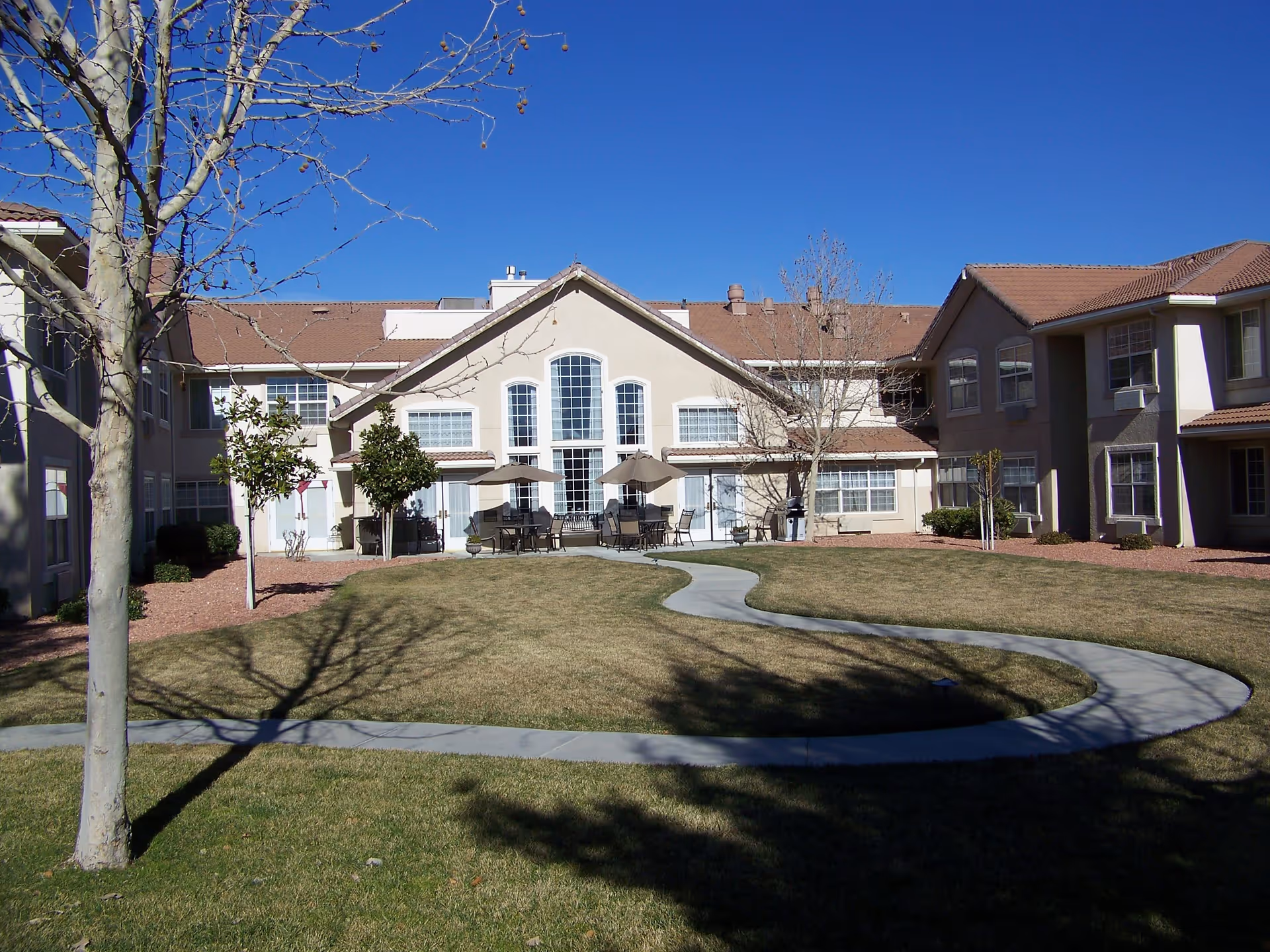 Outdoor view of a senior living facility with a curved concrete pathway leading through a grassy lawn towards a building with large windows and patio tables with umbrellas. Leafless trees and small shrubs are planted around the lawn under a clear blue sky.