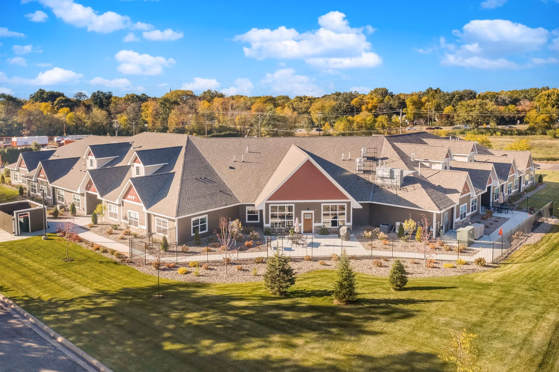 Aerial view of a single-story senior living facility building with multiple peaked roofs, surrounded by a well-maintained lawn and landscaping. The building has a combination of gray and red exterior walls with white trim. Trees and a clear blue sky with some clouds are visible in the background.