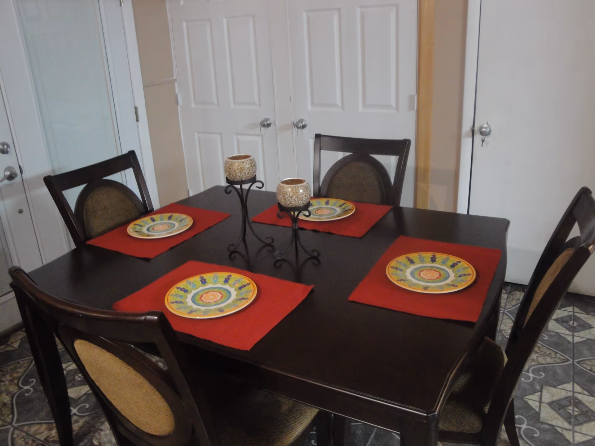 A dining room with a dark wooden table set for four people. Each place setting has a colorful plate on a red placemat. In the center of the table are two decorative candle holders with candles. The room has patterned tile flooring and white doors in the background.
