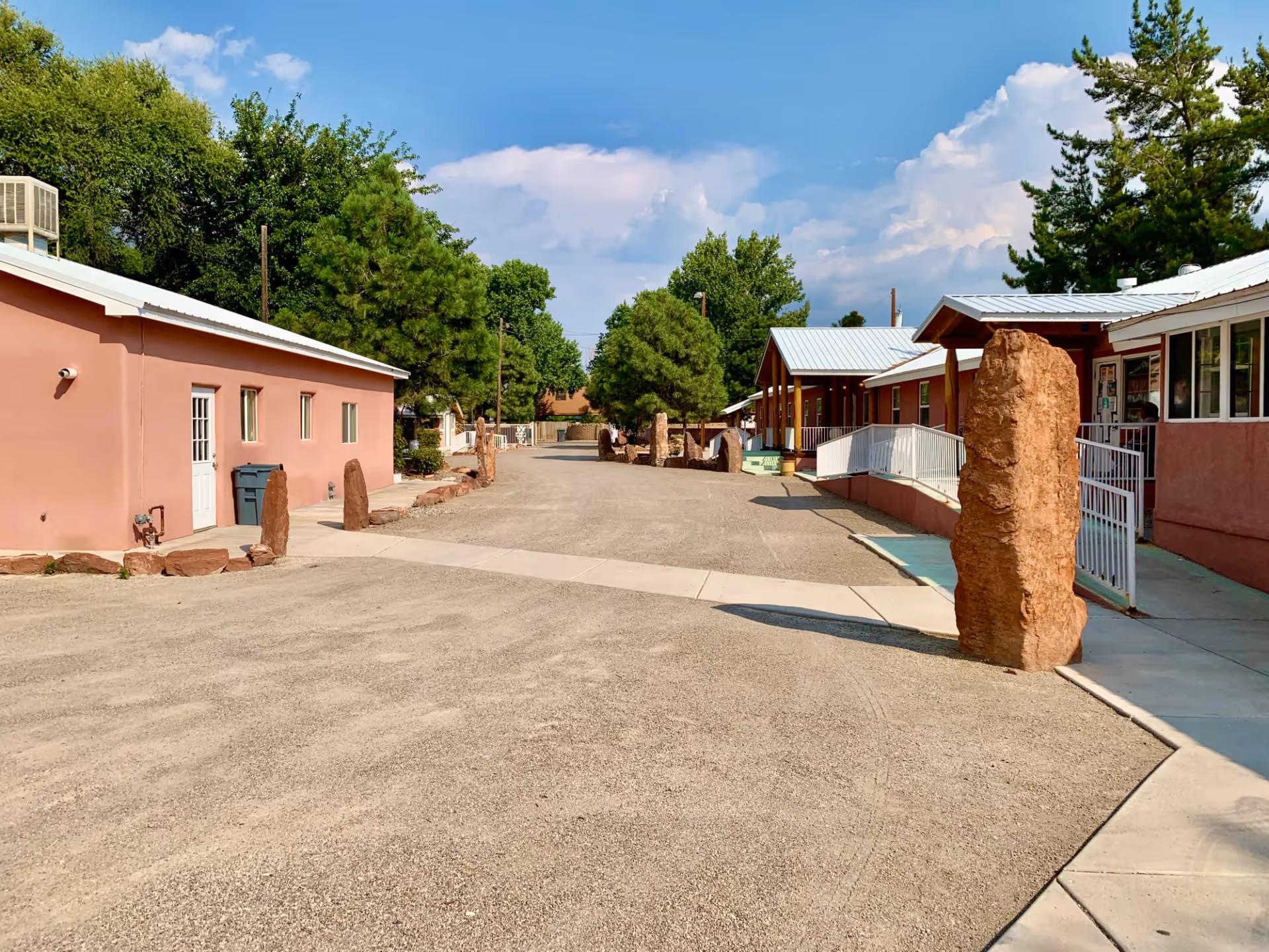 Outdoor view of Casa de Rosa Assisted Living facility showing a wide gravel pathway lined with tall stone pillars and bordered by single-story buildings with pinkish walls and white roofs. Trees and a partly cloudy blue sky are visible in the background.