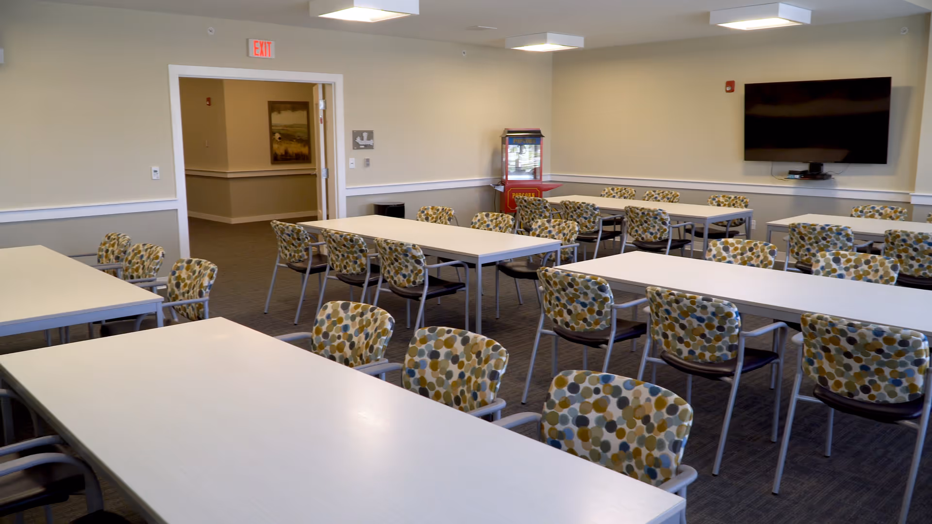 Well-lit communal dining/activity room with rows of white tables, patterned chairs, a wall-mounted TV, and a popcorn machine.