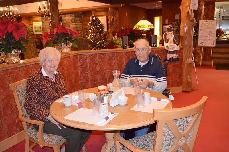 Two elderly individuals sitting at a round dining table set with placemats, glasses, cups, and condiments in a warmly decorated room with Christmas decorations including a Christmas tree and poinsettias.