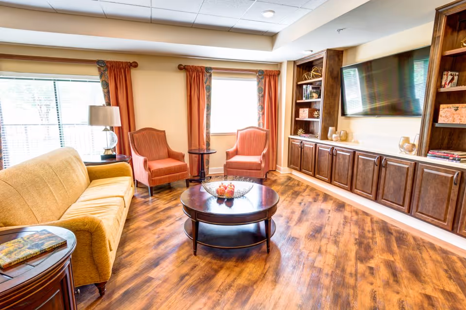 A bright and cozy living room with a yellow sofa, two orange armchairs, a round wooden coffee table with decorative items, wooden built-in shelves and cabinets, a large flat-screen TV mounted on the wall, and windows with orange curtains letting in natural light.