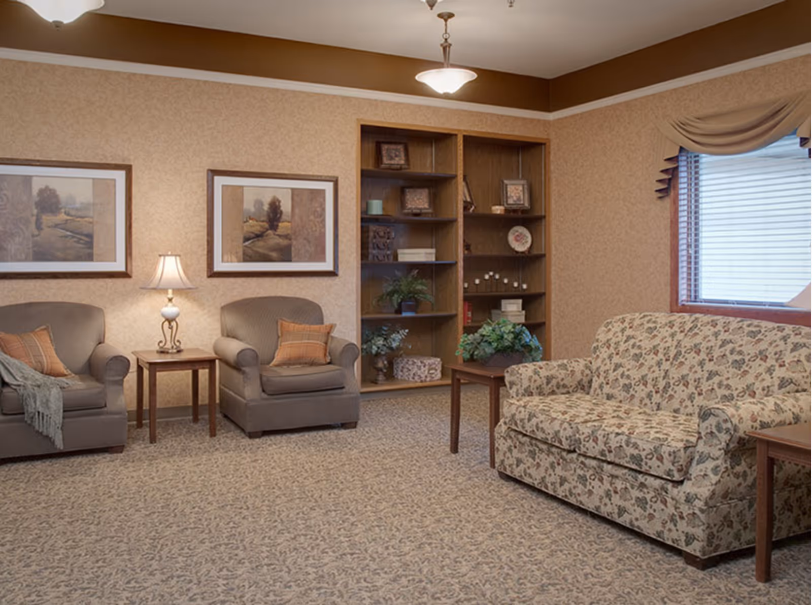 Warmly decorated sitting room with two upholstered armchairs, a floral sofa, side tables, a lamp, and a built-in bookshelf.